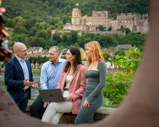 Vier Personen sitzen im Freien und arbeiten an einem Laptop, mit einer Burg im Hintergrund.